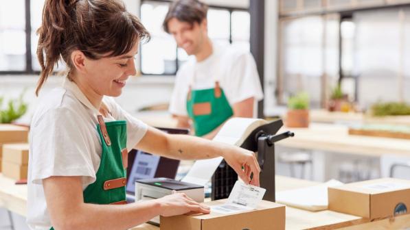 A woman wearing a green apron smiles while placing a shipping label on a package, with a coworker in the background.