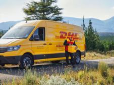 A DHL employee stands in front of a DHL delivery van on a country road, surrounded by green meadows and conifers with a mountain backdrop, checking a parcel.