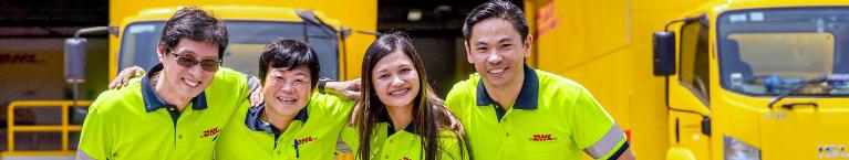 Four people in yellow DHL shirts in front of DHL vehicles smiling at the camera.