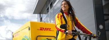 Eine lächelnde Zustellerin auf einem DHL-Fahrrad mit rot-gelber DHL-Jacke.