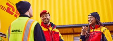 A smiling group of people, one female and two males, in DHL clothing in front of a yellow DHL building. 