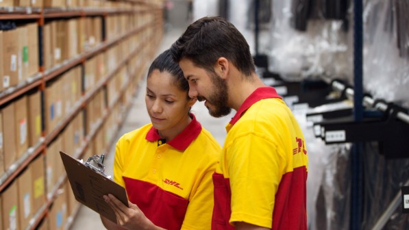 Zwei Kollegen in DHL-Kleidung schauen auf ein Klemmbrett, während sie in einem Lagerhaus stehen. 