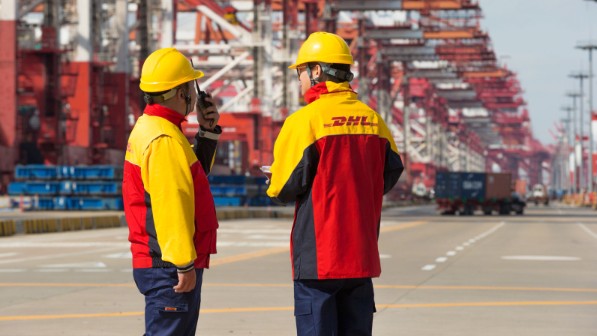 Two employees with red and yellow DHL jackets and yellow helmets at a transshipment site.