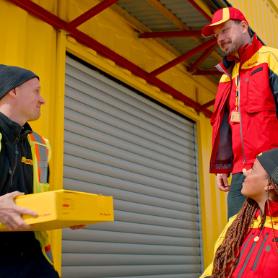 Zwei Männer und eine Frau in DHL-Kleidung vor einem DHL-Container.