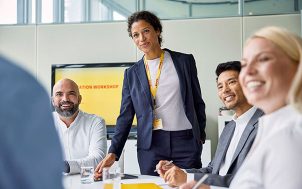 Two men and a woman sit at a conference table while another woman with a DHL lanyard is standing . 