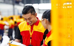 A man in DHL clothing and with a DHL lanyard smiles down on a parcel. 